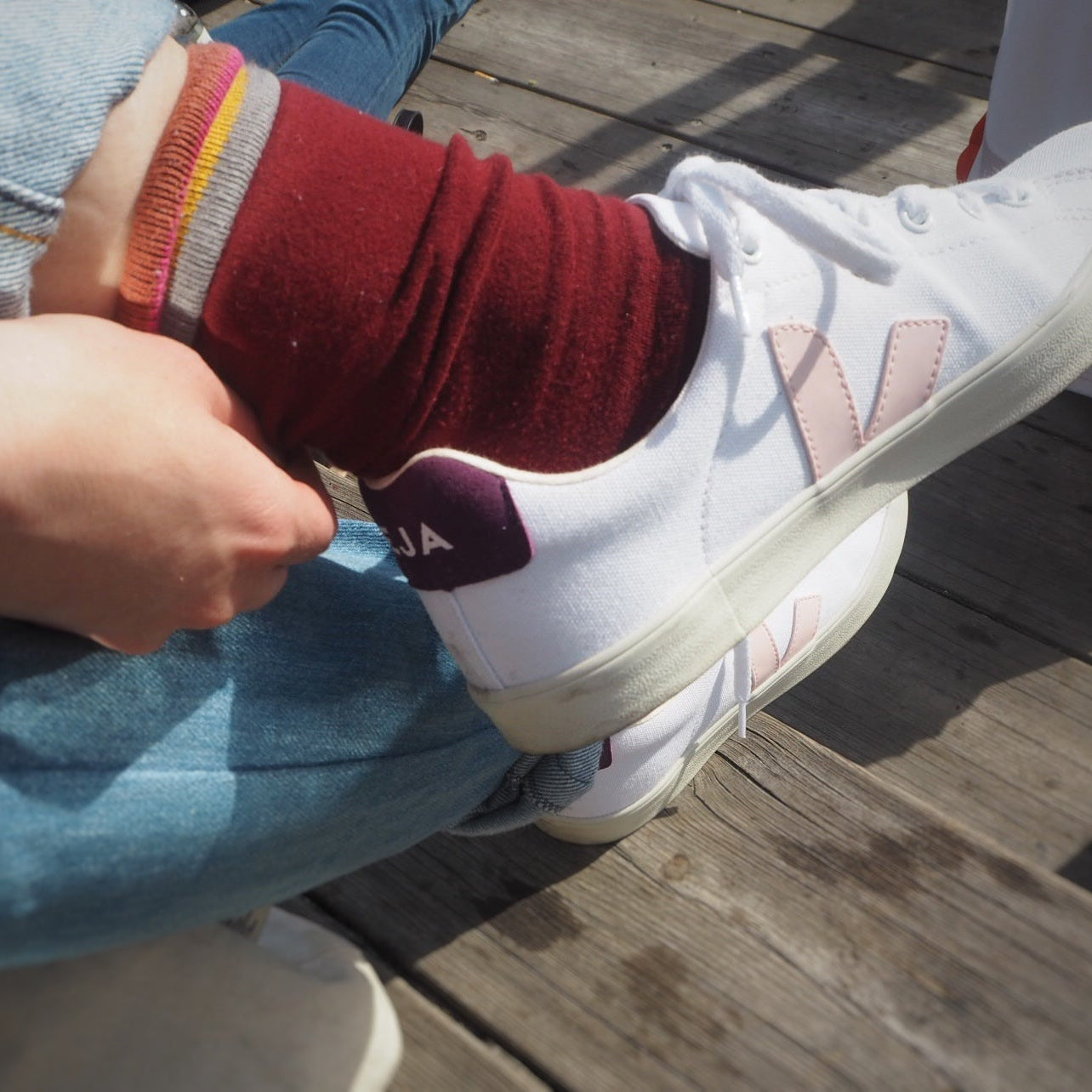 Person wearing a white sneaker with a brand logo, red sock, and blue jeans sitting on a wooden floor.